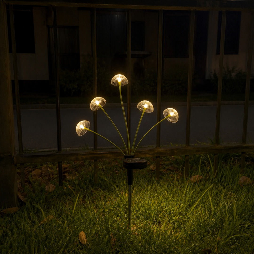 Antejardín de una casa chilena de villa, de noche, con una estaca de medusas encendida en el pasto junto a la reja